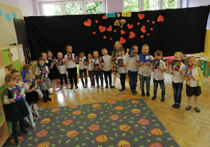 Zdjęcie dzieci z grupy fioletowej, dzieci stoją w kręgu z tyłu dekoracja z sercami dzieci ubrane są odświętnie prezentują kartki wykonane dla Mamy